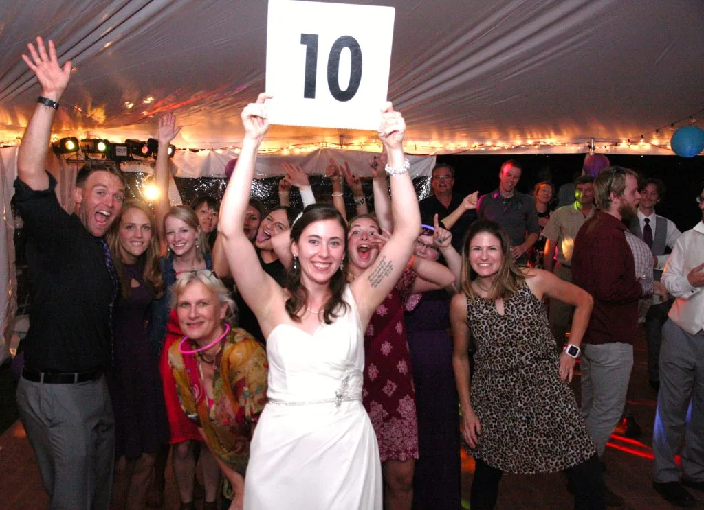 A bride holds up a '10' scorecard surrounded by cheering guests under string lights